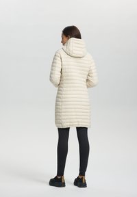 Gequilte crèmekleurige pufferjas met een capuchon, met een lengte tot halverwege de bovenbenen, textuur en lange mouwen, gecombineerd met zwarte legging en schoenen.
