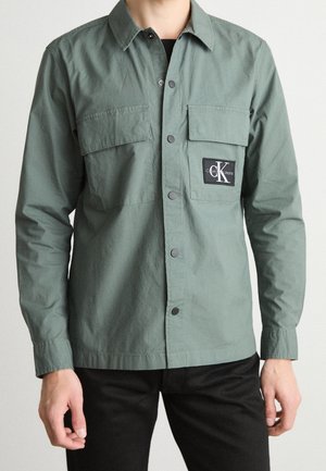 Hombre con camisa verde abotonada con dos bolsillos en el pecho, parche negro con el logo de Calvin Klein Jeans en el bolsillo derecho y pantalones negros.