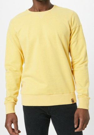 Mand iført en ensfarvet pastelgul langærmet sweater med ribstrikkede manchetter og kant, kombineret med sorte bukser.