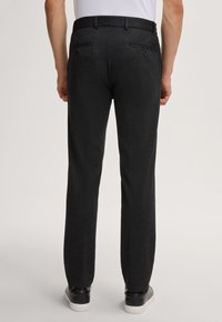 Uomo che indossa pantaloni grigi scuri a gamba dritta, sneakers nere con suole bianche e una camicia bianca, in piedi su uno sfondo neutro.