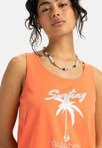 Kvinna som bär ett färgglatt pärlhalsband och en orange linne med vit palmträd och texten "Surfing Paradise".