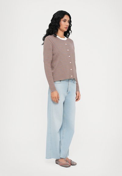Striped button-up cardigan in earthy tones, paired with wide-leg light blue jeans and multicolored flat shoes. Hair styled in loose waves.