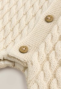 Beżowy sweter robiony na drutach z wzorem w warkocze, ozdobnymi brązowymi guzikami i prążkowanymi mankietami. Miękka tekstura i warstwowe detale w designie.