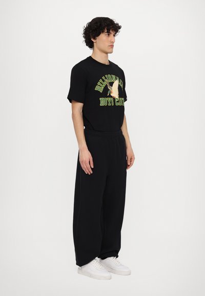 T-shirt graphique noire avec design vert et blanc, associée à un pantalon de jogging noir ample et des baskets blanches. Silhouette simple et décontractée.