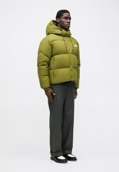 Giacca a vento verde con colletto alto, chiusura a zip, cordino regolabile. Abbinata a pantaloni grigi e scarpe in bianco e nero.