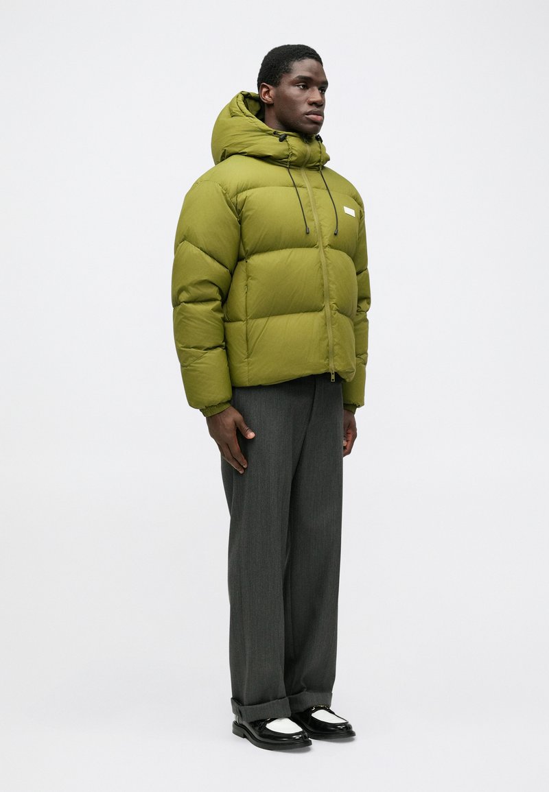 Green puffer jacket with a high collar, zip closure, adjustable drawstring. Paired with gray trousers and black-and-white shoes.