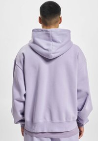 Haut de survêtement oversized violet clair avec capuche à cordon, épaules tombantes, ourlet côtelé et tissu texturé. La vue de dos montre une construction lisse.