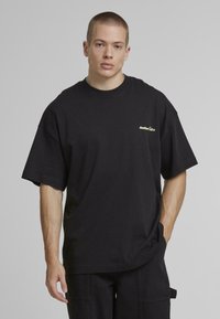 Tricou din bumbac negru, cu o croială lejeră, gât rotund și mâneci scurte. Prezintă un mic logo auriu pe piept.