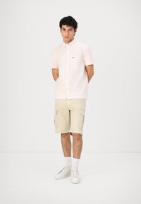 Camicetta rosa chiaro a maniche corte con colletto button-down, abbinata a pantaloni cargo beige e calzini bianchi. Sneakers bianche completano il look.