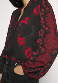 Czarna i czerwona bluzka damska z długimi rękawami, z przodu w stylu tie-dye i wzorzystymi rękawami, włożona w czarne dżinsy z czarnym paskiem, ręka w kieszeni.