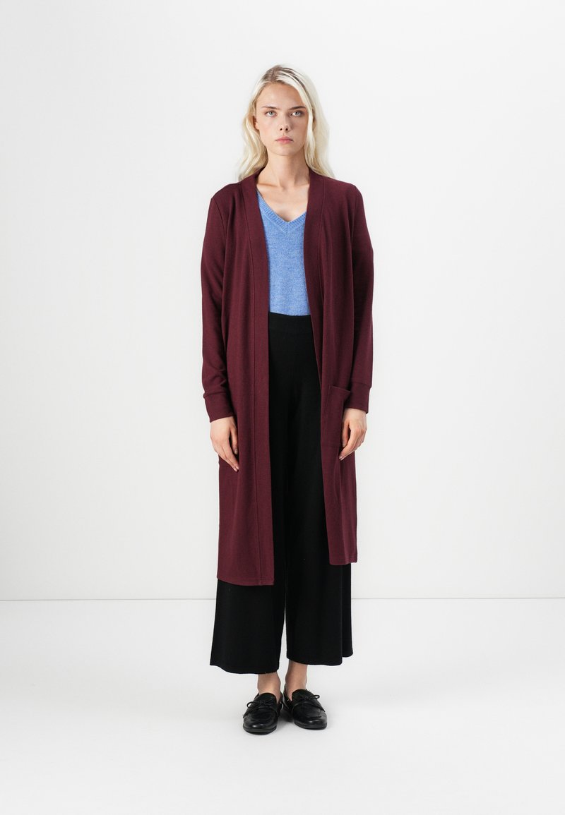 Bordeaux lange cardigan over een blauwe V-hals top, gecombineerd met zwarte wijde broek en zwarte schoenen. Glad textuur, open voorkant ontwerp.