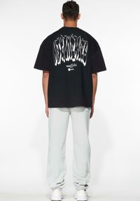 Schwarzes, übergroßes T-Shirt mit weißen stilisierten Text auf der Rückseite, kombiniert mit hellgrauen Hosen und weißen Turnschuhen. Einfaches Design, Baumwollmaterial.