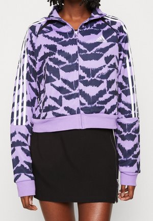 Žena nosí fialovo-černou bundu Adidas na zip se vzorem a bílými pruhy na rukávech a černou sukni se zipem na boku.