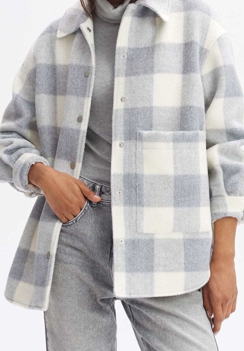 Chaqueta de franela a cuadros gris y blanco con cuello, botones a presión y un bolsillo frontal. Textura suave y corte relajado.