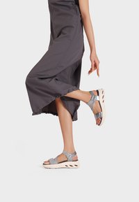 Abito di stoffa grigio con orlo a volant. La modella indossa sandali metallici argento con più cinturini e una suola arrotondata e imbottita.