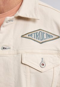 Chaqueta de mezclilla de tono beige claro con un logo bordado en forma de diamante en azul y dorado, un bolsillo en el pecho y detalles de costura en contraste.