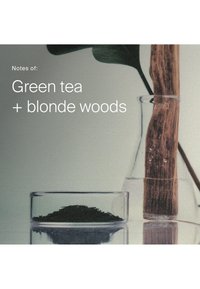 Glassbeholdere inneholder tørkede grønne teblader og et stykke lyst tre med et grønt blad i bakgrunnen. Teksten noterer duftdetaljer.