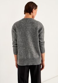 Vrouw met kort bruin haar, gekleed in een grijze gebreide trui en zwarte broek, met haar rug naar de camera gekeerd tegen een effen lichte achtergrond.