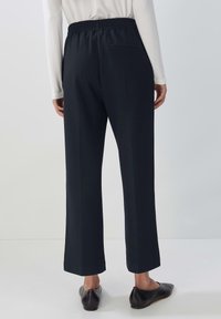 Kvinna som bär svarta vida byxor med elastisk midja, kombinerat med svarta lägenheter, stående mot en neutral bakgrund.