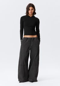 Sort langærmet top med lynlås, kombineret med løstsiddende, mørkegrå bukser med vide ben, elastik i taljen og synlige syningsdetaljer.