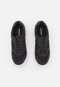 Ett par svarta mesh och lädersneakers med vita sulor och svart-vita snören, visade uppifrån på en vit bakgrund.