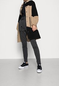 Veste surdimensionnée deux tons beige et noire avec des manches larges, associée à un pantalon gris ajusté et des baskets noires avec semelles blanches.