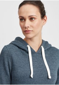 Hooded mikina tmavomodrej farby, so bielymi šnúrkami a mäkkou textúrou. Dizajn je ležérny s voľným strihom.