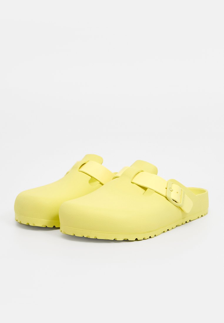 Gelbe Clogs mit einer glatten, matten Oberfläche; breite, abgerundete Zehenpartie; einziger verstellbarer Riemen; strukturierte Sohle für besseren Halt.