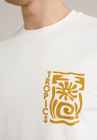 Balta kokvilnas T-kreklu ar zelta grafisku izdruku, kurā ir vārds "TROPICS", stilizēts zieds un "svārka" raksti kreisajā krūšu daļā.