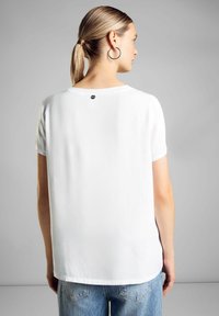 T-shirt bianca a maniche corte realizzata in tessuto liscio, con un delicato dettaglio del logo sul retro e una vestibilità comoda, abbinata a jeans blu.