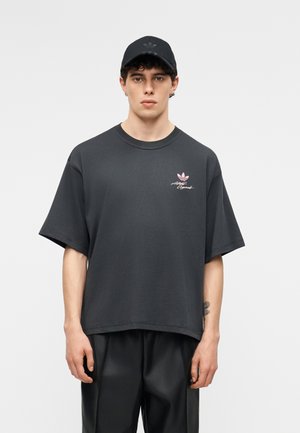 Νέος άνδρας φορώντας μαύρο καπέλο Adidas και υπερμεγέθη σκούρα γκρι μπλουζάκι με ροζ λογότυπο στο στήθος, στέκεται μπροστά σε απλό φωτεινό φόντο.