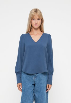 Blå langermet bluse med V-hals og avslappet passform; har puffermer og myk, teksturert stoff; kombinert med lyseblå jeans.