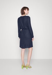 Vestido azul-marinho até ao joelho com mangas compridas e tecido texturizado, cinto na cintura. Usado com sandálias de salto alto verdes.