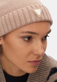 Berretto beige lavorato a maglia con tessuto a coste e una toppa triangolare con logo. Vista ravvicinata che mette in evidenza il tessuto morbido e il design strutturato.