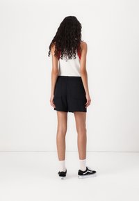 Černé kraťasy s uvolněným střihem, elastickým pasem, dvěma zadními kapsami, nošeny s bílým tílkem a černými teniskami. Viditelné jsou kudrnaté vlasy.