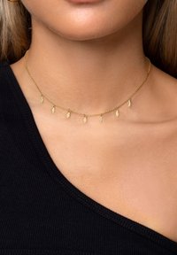 Collana choker in oro con piccoli charm a forma di foglia distribuiti uniformemente lungo una delicate catena, indossata su un tessuto nero per il contrasto.