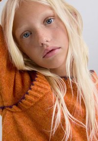 Suéter naranja de punto con acentos morados, que presenta un cuello acanalado y textura. Cabello rubio largo y liso enmarca el rostro con ojos azules.