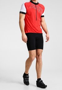 Jersey de ciclismo rojo y blanco con media cremallera, que presenta un patrón geométrico. Lleva puesto con pantalones cortos de ciclismo negros y zapatos negros.