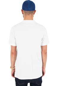 Tricou alb cu mânecă scurtă, cu croială dreaptă, textură netedă și tiv rotunjit, purtat cu blugi negri și o șapcă din plasă albastră.
