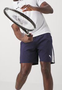 Polo blanc, associé à un short marine avec des accents blancs et un logo, tenu par une personne avec une raquette de tennis en pose active.