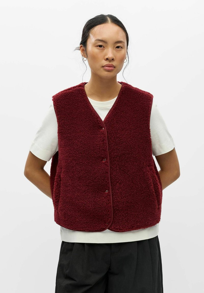 Femme aux cheveux foncés portant un gilet polaire bordeaux sans manches sur un t-shirt blanc à manches courtes et un pantalon noir, debout devant un fond uni.