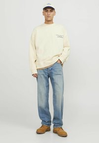 Cream sweatshirt with printed text, light blue jeans, and tan boots. Baseball cap with blue accents. Casual fit.
