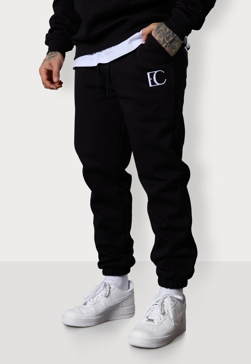 Schwarze Baumwoll-Jogginghosen mit elastischem Bund und Bündchen, versehen mit einem weißen gestickten Logo am linken Oberschenkel. Kombiniert mit weißen Sneakers.