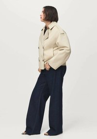 Beige dubbelknäppt jacka med stora knappar, sidofickor och platt krage, i kombination med vida mörka jeansbyxor och svarta sandaler.