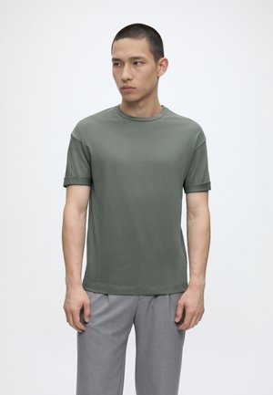 Jonge man met kort haar, een groen T-shirt met opgerolde mouwen en lichtgrijze broek, kijkend naar de zijkant tegen een effen achtergrond.