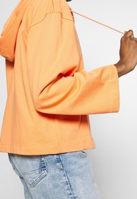 Rennokki oranssi huppari pehmeässä kankaassa, jossa on nyöri, leveät hihat ja lyhyt helma. Yhdistetty vaaleansinisiin farkkuihin.