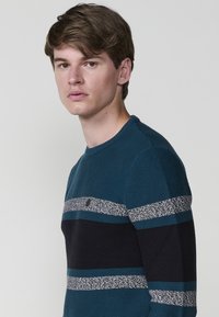 Jersey de color azul verdoso con patrones de rayas horizontales en azul verdoso más oscuro, gris y negro. Tejido ligero, escote redondo y puños acanalados.
