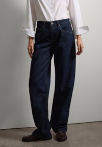 Jeans de mezclilla azul oscuro con un corte relajado, que presentan bolsillos frontales y costuras amarillas visibles. Se llevan con una camisa blanca de cuello.