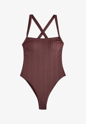 Jeune femme aux longs cheveux foncés portant un maillot de bain une pièce marron, debout près d'une rambarde au bord de la mer sous un ciel nuageux.
