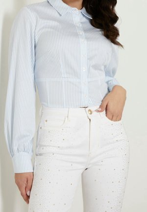Femme portant une chemise à rayures bleu clair et blanc boutonnée et un jean blanc avec de petits clous dorés en décoration.